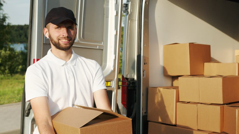 A young man with a beard, wearing a white polo shirt and a black cap, is standing outside next to an open vehicle, possibly a removal van, during daylight. He is holding a medium-sized cardboard box with both hands, preparing for a furniture transport or home relocation process. The van's interior is visible behind him, showing a partially open door and part of the vehicle’s structure. Inside the van, there are several stacked cardboard boxes and plastic-wrapped items, indicating packing for a move. The surrounding environment features greenery and a clear sky, suggesting an outdoor loading area. Tooting Man and Van’s home relocation and removals services are implied by the scene, reflecting the logistics involved in packing, loading, and transport of household items, with appropriate labeling of tools such as cardboard boxes and the van used in professional removals.