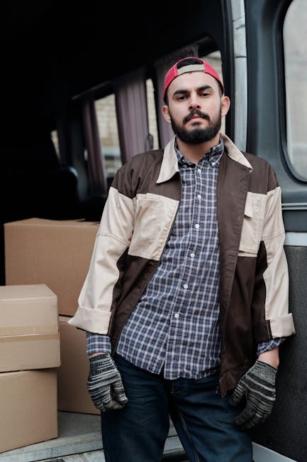 A man with a beard, wearing a pink cap, a checked shirt, and a beige and brown jacket, stands inside a vehicle used for house removals, with one hand in his pocket and the other resting on a box. Behind him, there are several cardboard boxes stacked on the vehicle's floor, ready for transportation during a home relocation. The interior of the van features a dark ceiling and fabric-lined walls, illuminated by natural light coming through the side windows. The scene captures a loading process typical of furniture transport and packing and moving activities, with protective blankets, straps, and other equipment possibly visible in the background. This image exemplifies professional removal services provided by Tooting Man and Van, specifically focusing on house removals near Tooting Bec Lido, illustrating the preparation and logistics involved in a house move.