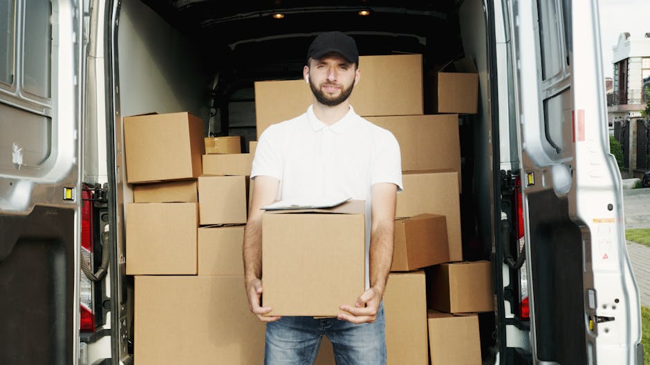 A young man with a beard, wearing a white polo shirt and a black cap, is standing outside next to an open vehicle, possibly a removal van, during daylight. He is holding a medium-sized cardboard box with both hands, preparing for a furniture transport or home relocation process. The van's interior is visible behind him, showing a partially open door and part of the vehicle’s structure. Inside the van, there are several stacked cardboard boxes and plastic-wrapped items, indicating packing for a move. The surrounding environment features greenery and a clear sky, suggesting an outdoor loading area. Tooting Man and Van’s home relocation and removals services are implied by the scene, reflecting the logistics involved in packing, loading, and transport of household items, with appropriate labeling of tools such as cardboard boxes and the van used in professional removals.
