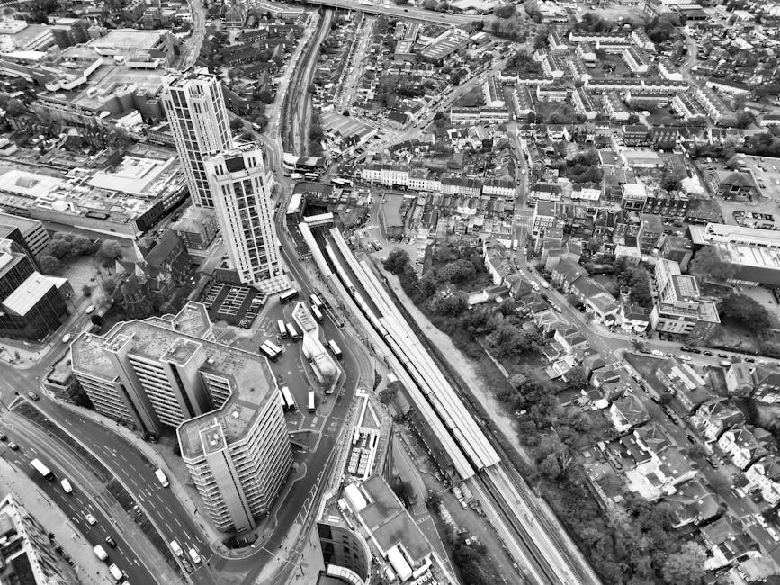 An aerial black and white photograph shows a dense urban area with high-rise and mid-rise buildings, streets filled with traffic, and a railway station with train platforms and tracks extending through the scene. The surrounding infrastructure includes roads, bridges, and parking areas. The image captures the busy environment of a cityscape, likely within central London, near Tooting Broadway station in the SW17 district. This setting relates to the services offered by Tooting Man and Van, who specialise in removals, house relocations, and furniture transport, with a focus on efficient loading and moving logistics within urban environments.
